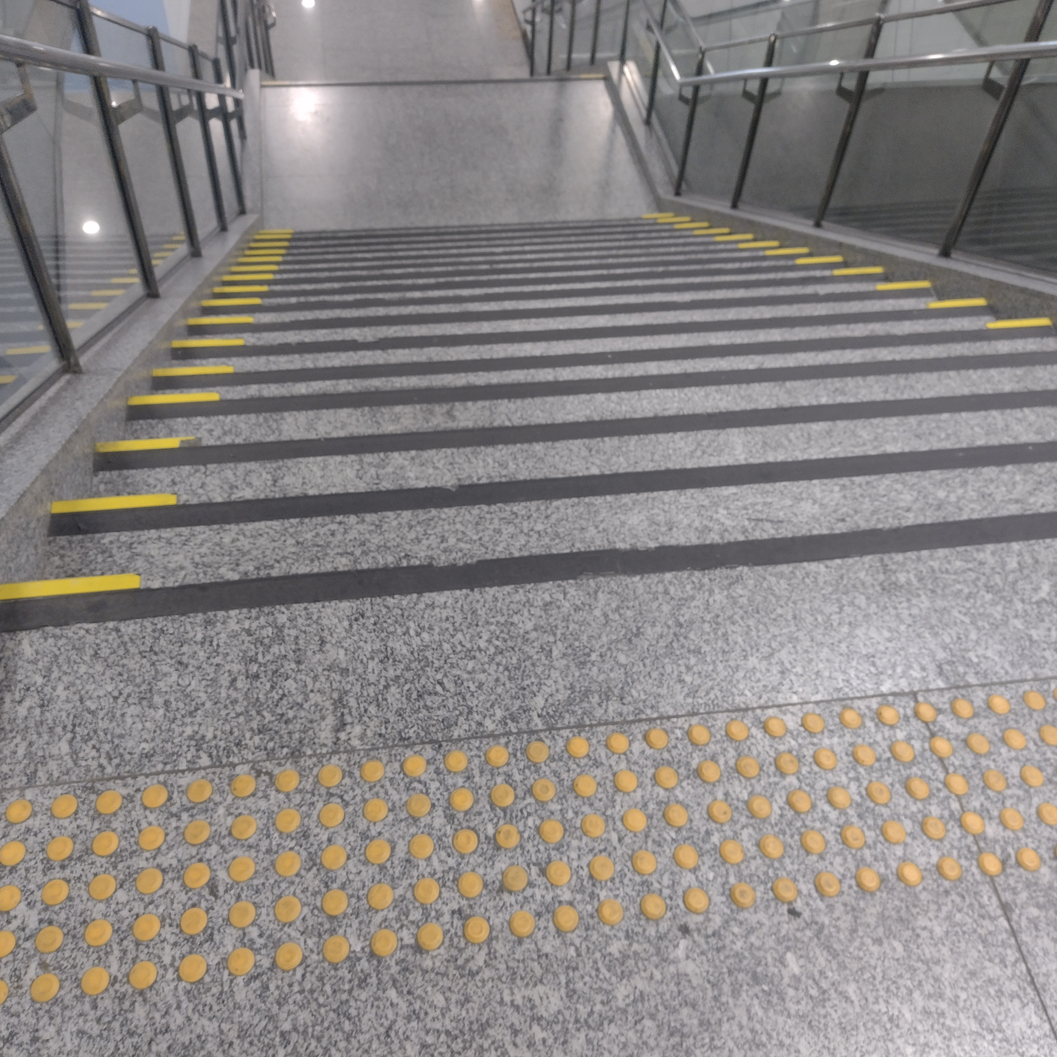 Escadaria com piso de granito e sinalização tátil de alerta na área de aproximação. Os degraus possuem sinalização visual de contraste nas bordas e há corrimãos e guarda-corpos laterais em vidro e metal.
