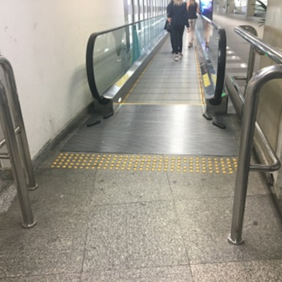 Entrada de uma esteira rolante com piso tátil em frente à sua base e corrimãos laterais. Há sinalização em amarelo indicando a presença do equipamento.