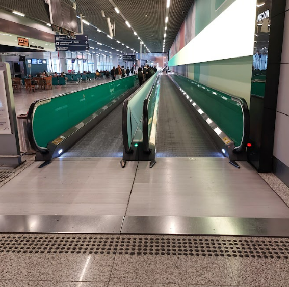 Esteira rolante ampla, com entrada nivelada e corrimãos verdes dos dois lados. O piso tem textura e sinalização tátil na área de embarque.