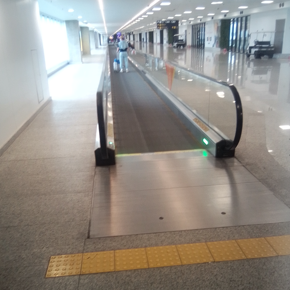 Esteira rolante em aeroporto com corrimãos de vidro e sinalização verde de funcionamento. Há piso tátil na entrada para orientação de pessoas com deficiência visual.