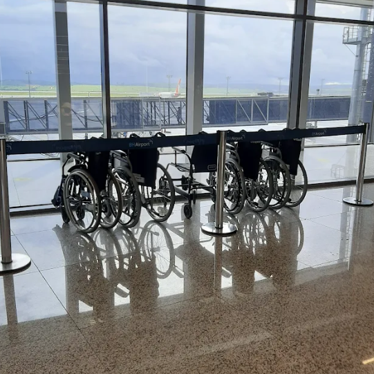Conjunto de cadeiras de rodas manuais enfileiradas e organizadas atrás de divisórias no saguão do aeroporto BH Airport, com fundo envidraçado mostrando o pátio de aeronaves.
