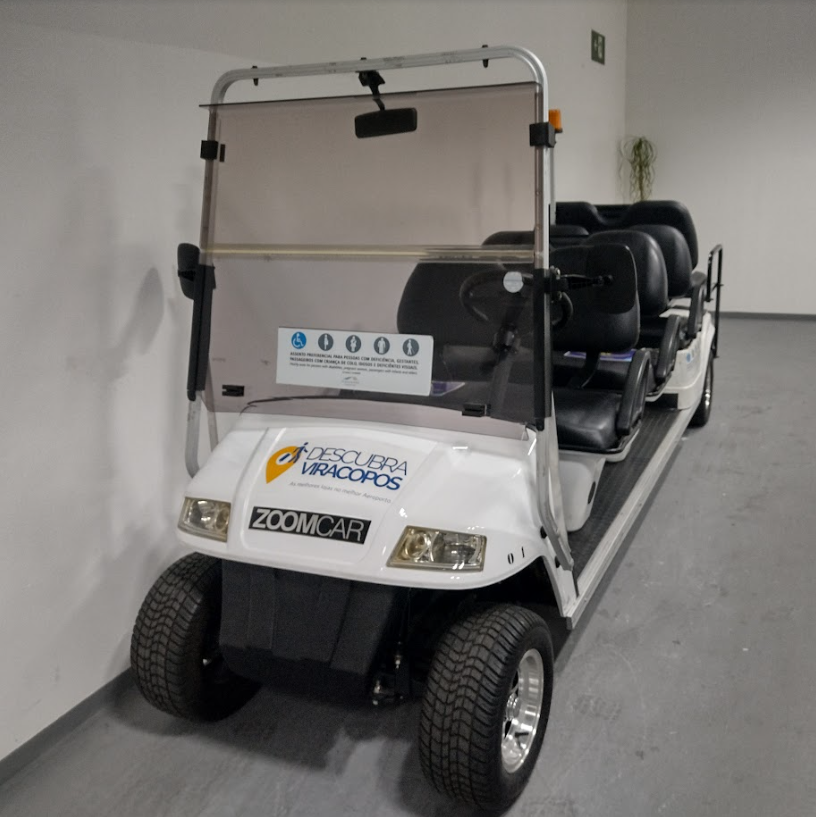 Carrinho elétrico branco de transporte de passageiros no Aeroporto de Viracopos, com quatro fileiras de assentos e identificação "Zoomcar" na frente. Há um adesivo informando o uso prioritário para pessoas com deficiência, mobilidade reduzida, gestantes e idosos.