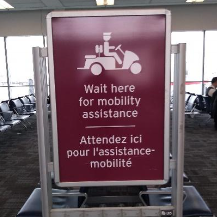 Placa de cor vinho posicionada entre assentos de sala de embarque com a mensagem em inglês e francês: “Wait here for mobility assistance / Attendez ici pour l'assistance-mobilité”, indicando ponto de espera para auxílio de transporte.