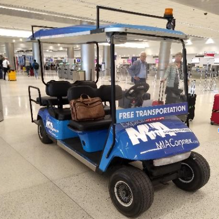 Carrinho elétrico azul identificado com a inscrição “FREE TRANSPORTATION” e o logotipo “MIAmover MIA ConnecTion” estacionado dentro do aeroporto, pronto para transportar passageiros, com assentos e bagageiro dianteiro.
