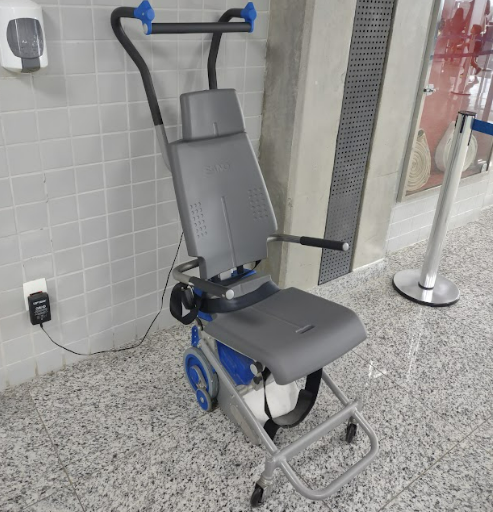 Cadeira especial motorizada com trilhos de borracha e apoio de braço para auxiliar no transporte de pessoas com deficiência em escadas. Está posicionada em área interna com piso de granito e parede de azulejo claro.