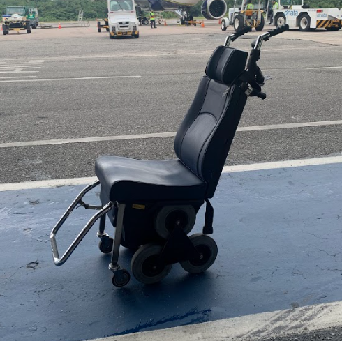 Cadeira adaptada com rodas em estrela para permitir a subida e descida de escadas, localizada na pista do aeroporto, ao lado de uma aeronave.