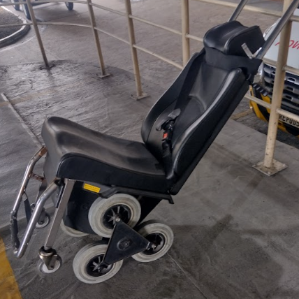 Cadeira com o mesmo modelo da anterior, projetada para subir escadas, posicionada ao lado de um corrimão em área coberta do aeroporto, com piso de concreto.