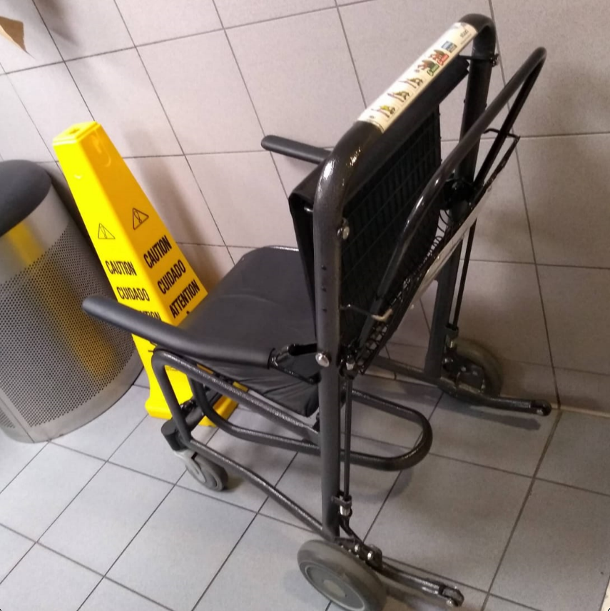 Cadeira de rodas dobrável preta com estrutura metálica posicionada próxima a um cone de sinalização amarelo em ambiente interno, sugerindo uso para apoio na locomoção em espaços de acesso restrito.