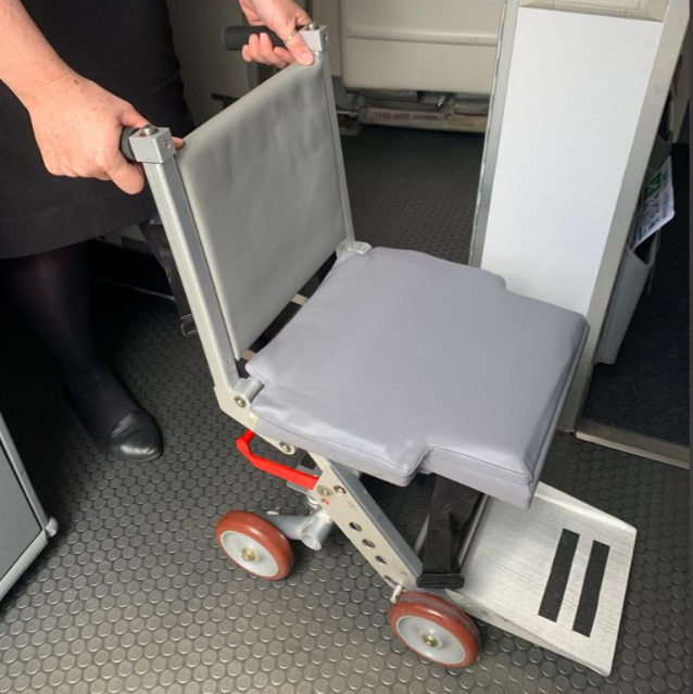 Cadeira de rodas estreita com assento cinza, alça para empurrar e quatro rodas pequenas, utilizada para locomoção de passageiros com mobilidade reduzida no interior de aeronaves.