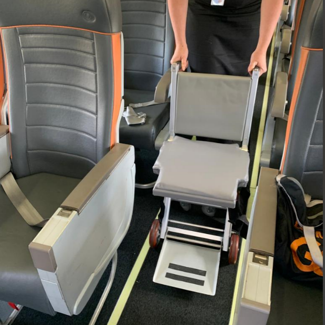 Cadeira de rodas estreita adaptada posicionada entre fileiras de poltronas de avião, demonstrando acessibilidade para embarque e acomodação no interior da cabine