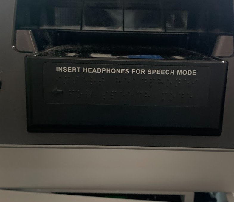Detalhe do aviso em braile e texto em alto-relevo sobre o acionamento do modo por voz ao inserir fones de ouvido, presente em um totem de autoatendimento.
