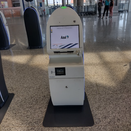Totem de autoatendimento da companhia Azul posicionado em área ampla, com tela acessível, estrutura compacta e espaço livre abaixo que permite aproximação frontal por usuário em cadeira de rodas.