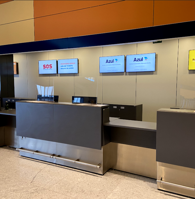 Balcões de atendimento de uma companhia aérea (Azul) em um aeroporto. Os balcões são cinza e prata, com monitores acima exibindo informações e o logo da empresa. Parte do balcão à esquerda é mais baixa, indicando um ponto de atendimento acessível. Ao fundo, a parede tem painéis em tons de laranja e bege.