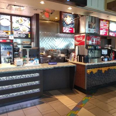 Balcão de atendimento de uma lanchonete ou restaurante fast-food. O balcão possui uma seção mais baixa à esquerda, projetada para acessibilidade. Ao fundo, veem-se menus iluminados, equipamentos de cozinha e máquinas de refrigerante.