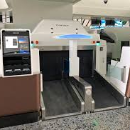 Equipamento de autoatendimento para despacho de bagagem em um aeroporto, com duas esteiras paralelas e um totem com tela à esquerda. A máquina é predominantemente branca com detalhes em marrom e luzes azuis.