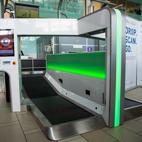 Equipamento de autoatendimento para despacho de bagagem, moderno e com design em tons de branco e cinza. Uma esteira está visível na parte inferior. Uma tela de autoatendimento está à esquerda e uma parede com iluminação verde destaca a área de despacho. Ao fundo, um banner com a inscrição "DROP. SCAN. GO." é parcialmente visível.