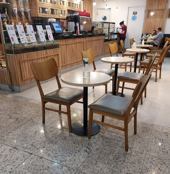 Interior de uma cafeteria ou lanchonete com mesas redondas e cadeiras de madeira. Ao fundo, um balcão de atendimento e prateleiras com produtos. O piso é de granito claro.