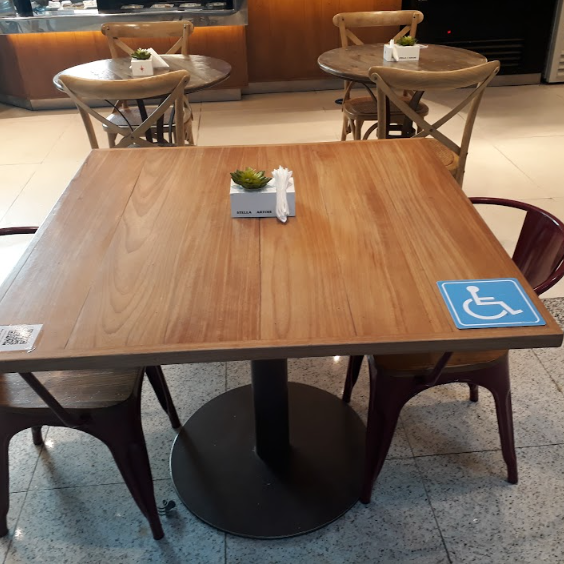 Mesa quadrada de madeira em uma área de alimentação, com cadeiras ao redor. Sobre a mesa, há um guardanapo e um pequeno vaso com uma planta. No canto direito da mesa, 