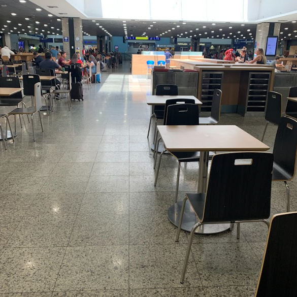 Ampla área de alimentação de um aeroporto com diversas mesas quadradas e cadeiras pretas e cinzas. O ambiente é bem iluminado por luz natural e artificial, com um piso claro. Ao fundo, pessoas circulam e outras estão sentadas às mesas.