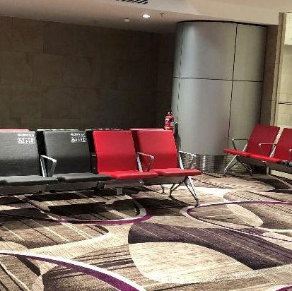 Fileira de assentos em uma sala de espera de aeroporto, com alguns assentos em vermelho e outros em cinza escuro, em um ambiente com piso acarpetado com padrões geométricos.