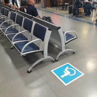 Fileira de assentos azuis em uma área de espera de aeroporto com piso cinza claro. Em primeiro plano, no chão, um adesivo azul e branco com o símbolo internacional de acesso indicando um espaço reservado.