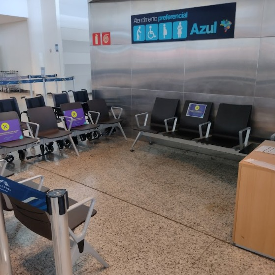 Área de espera em um aeroporto com diversos assentos. Alguns assentos possuem um aviso amarelo com um "X" roxo, indicando que não devem ser utilizados, possivelmente para distanciamento social. Ao fundo, uma placa sinaliza "Atendimento preferencial" com símbolos de acessibilidade e o logo da companhia aérea Azul.