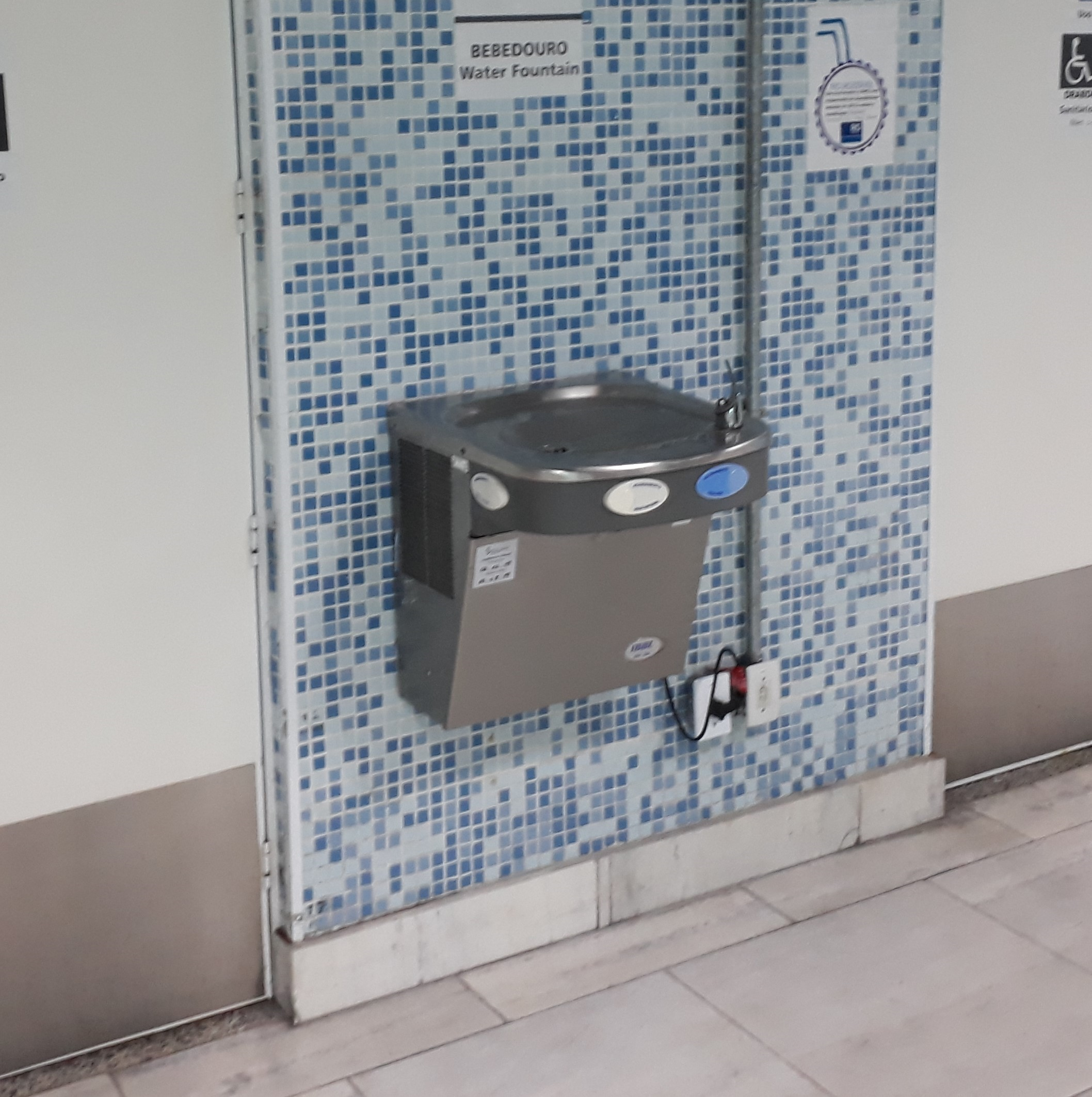 Bebedouro metálico instalado em parede com revestimento de pastilhas azuis, com altura acessível e espaço livre embaixo, permitindo o uso por pessoas em cadeira de rodas.