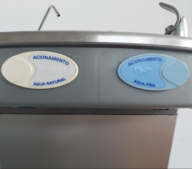 Close dos botões de acionamento de um bebedouro acessível, com identificação em Braille e indicação de água natural e água fria, demonstrando acessibilidade sensorial.