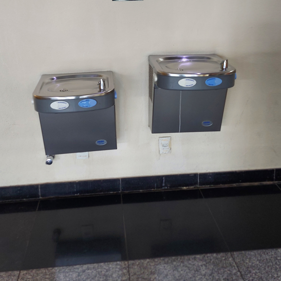 Dois bebedouros instalados lado a lado em parede clara, com alturas diferentes, sendo um deles mais baixo para permitir o uso por pessoas em cadeira de rodas. Ambos possuem botões acessíveis com identificação em Braille.