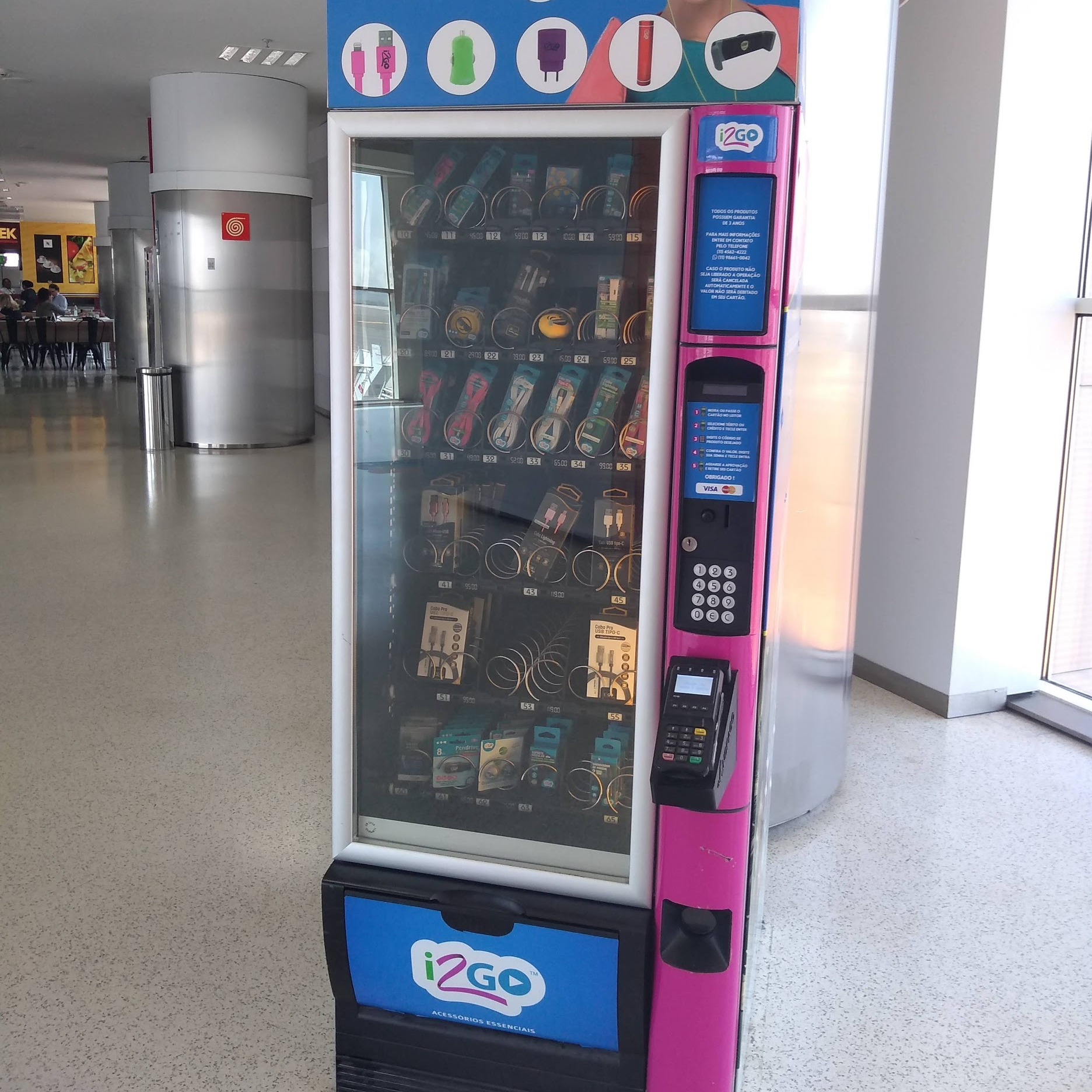 Máquina de venda automática localizada no saguão de um aeroporto, com produtos eletrônicos como cabos e carregadores, teclado numérico acessível, tela e leitor de cartão posicionados em altura reduzida.