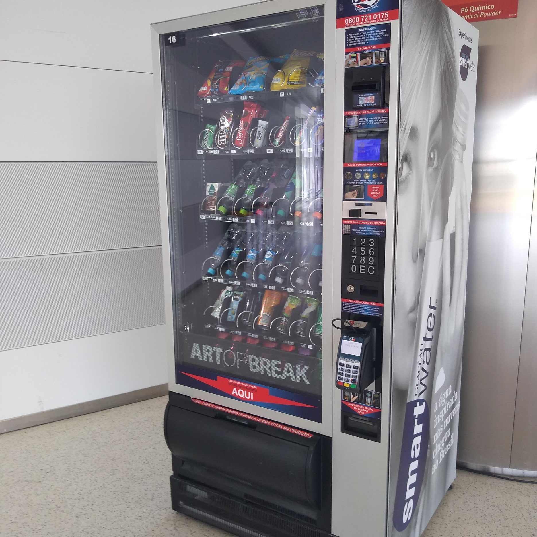 Máquina de venda automática de alimentos e bebidas com tela e teclas posicionadas de forma acessível. O equipamento oferece opções diversas e possui terminal de pagamento acessível.