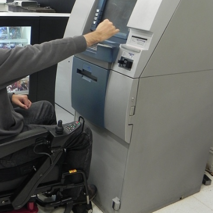 Pessoa utilizando cadeira de rodas interage com um caixa eletrônico. A tela, teclado e leitor de cartão estão posicionados em altura apropriada para acesso frontal.