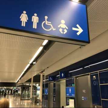 Placa suspensa azul com pictogramas indicando localização de sanitários masculino, feminino, acessível e fraldário.