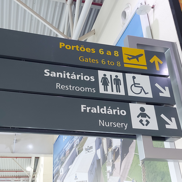 Sinalização suspensa com setas indicando o caminho para sanitários (incluindo acessível) e fraldário, com pictogramas e palavras em português e inglês.