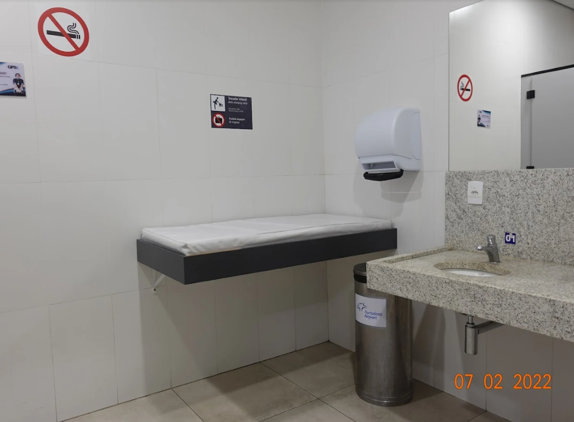 Espaço amplo com bancada de granito, pia acessível e trocador infantil fixado na parede, com almofada acolchoada e lixeira abaixo da pia. Placa de acessibilidade e data visível na imagem.