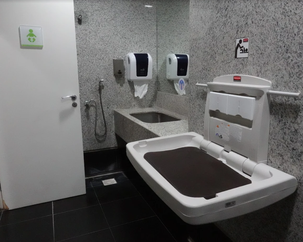 Ambiente com trocador de parede rebatível aberto, pia de apoio ao fundo e diversos dispensadores fixados na parede. Sinalização do trocador e símbolo de troca de fraldas na porta.