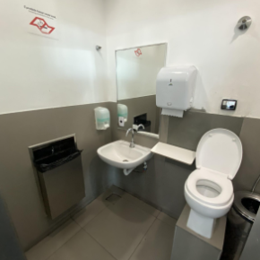 Ambiente de sanitário ostomizado com vaso sanitário elevado, pia ao lado com torneira, espelho e dispensadores de sabonete líquido e papel toalha, todos acessíveis.