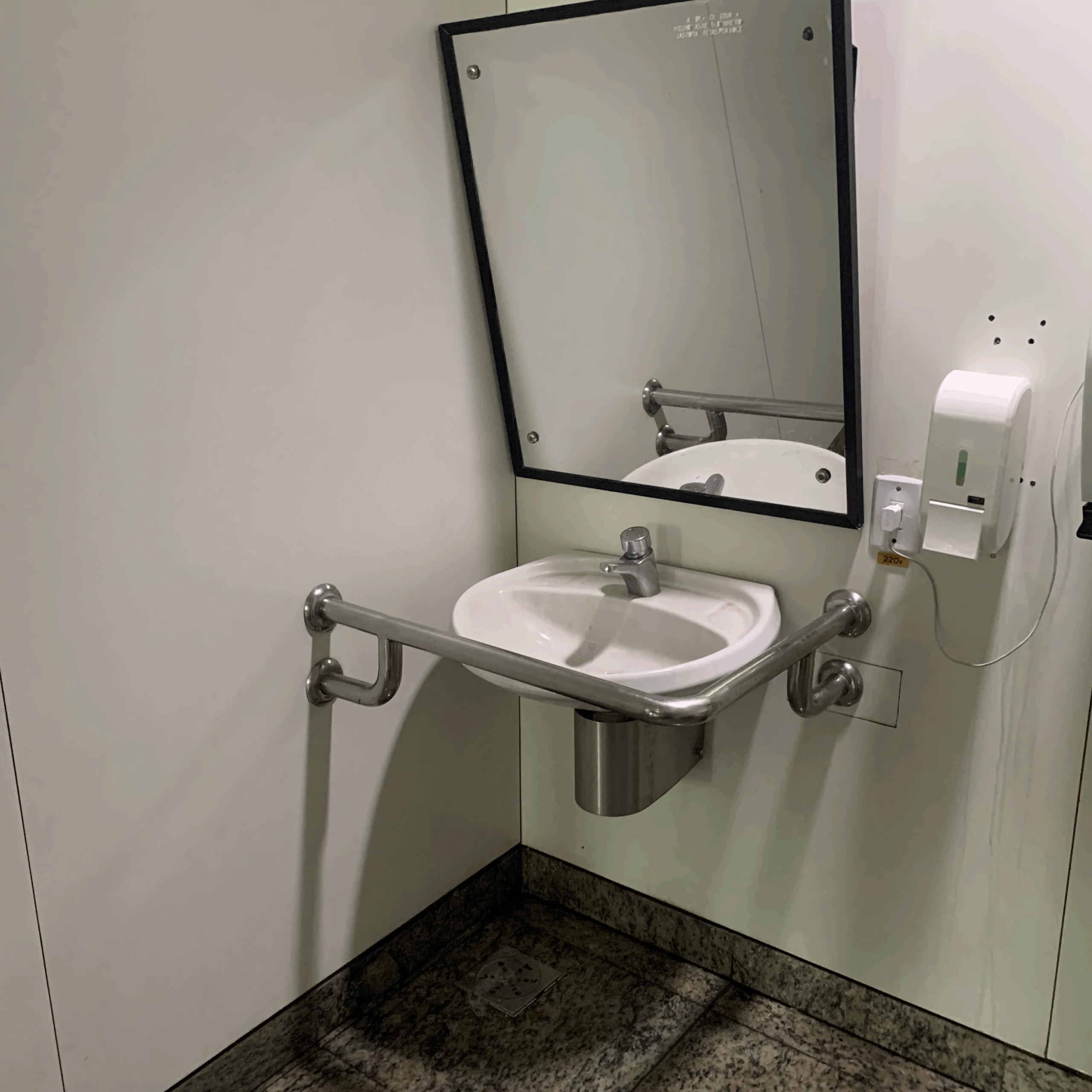 Lavatório acessível com espelho inclinado, barras de apoio metálicas ao redor da pia e dispenser de sabão líquido na parede lateral.