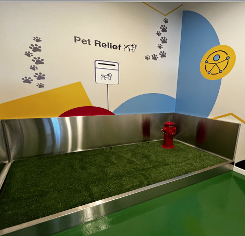 Área interna com piso de grama sintética cercado por estrutura metálica, sinalizada com “Pet Relief” e ícones de patas e cão, em parede colorida com símbolo de acessibilidade.