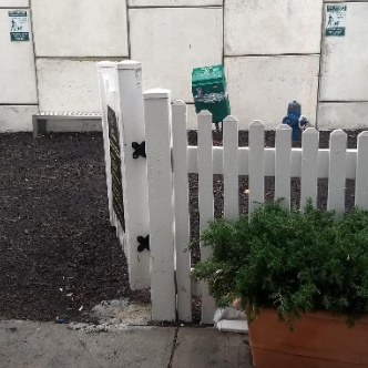 Espaço externo com cercado branco de madeira, poste higiênico e lixeira, sinalizado com placas verdes para uso de cães.