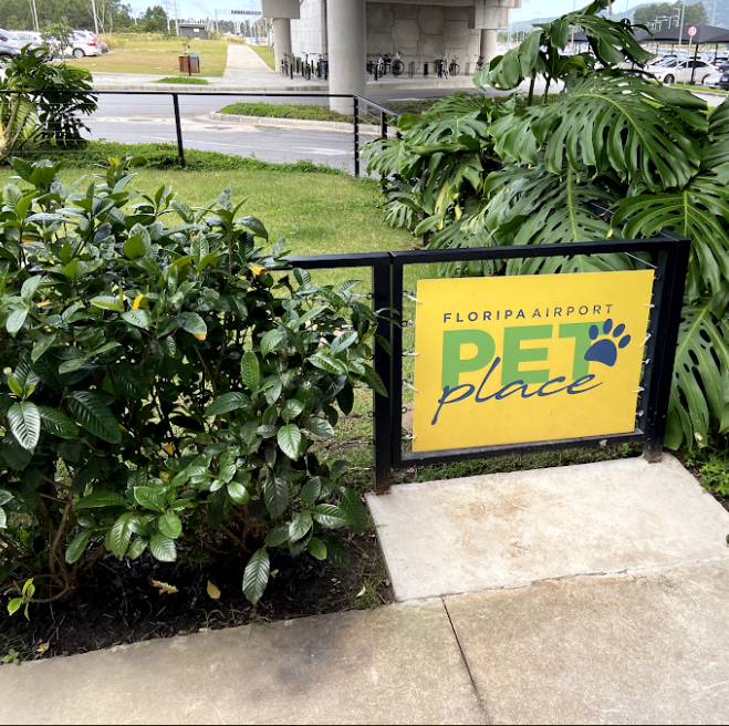 Entrada de área externa sinalizada como “Floripa Airport Pet Place”, com cercado preto, calçada acessível e vegetação ao redor.