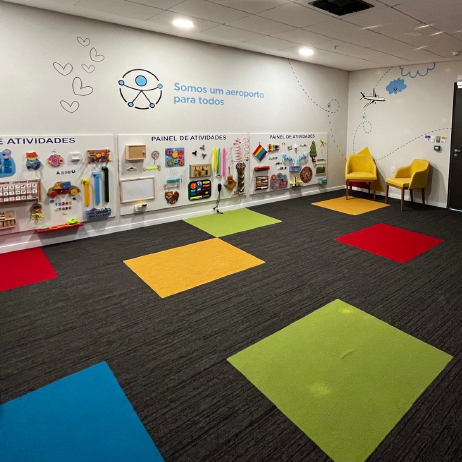 Sala sensorial com piso acarpetado e tapetes coloridos quadrados, equipada com painéis interativos de atividades fixados na parede, duas cadeiras amarelas e decoração lúdica com o texto "Somos um aeroporto para todos".