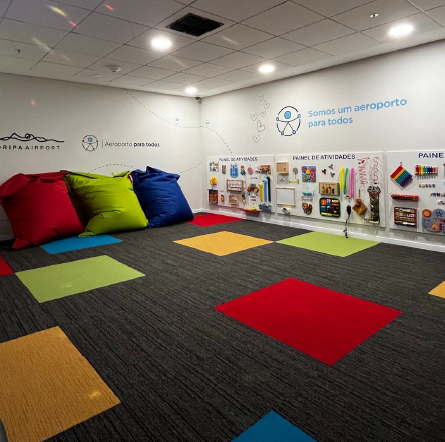 Ambiente sensorial com painéis táteis e interativos nas paredes, pufes coloridos no chão e tapetes em cores vibrantes, projetado para estimular e acalmar crianças em ambiente aeroportuário.