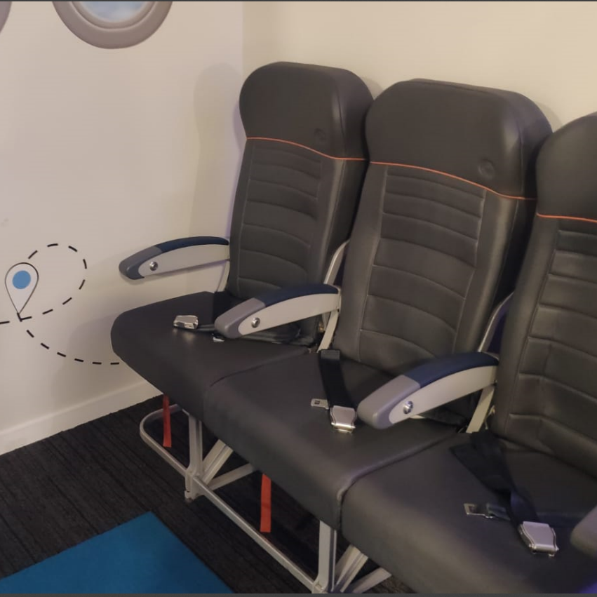 Três assentos simulando poltronas de avião, em sala sensorial com parede decorada com janelas fictícias e linha pontilhada representando trajeto, permitindo ambientação prévia de passageiros com deficiência.