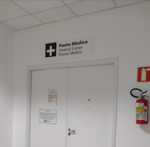 Entrada de posto médico com sinalização trilíngue (português, inglês e espanhol) e símbolo de cruz.